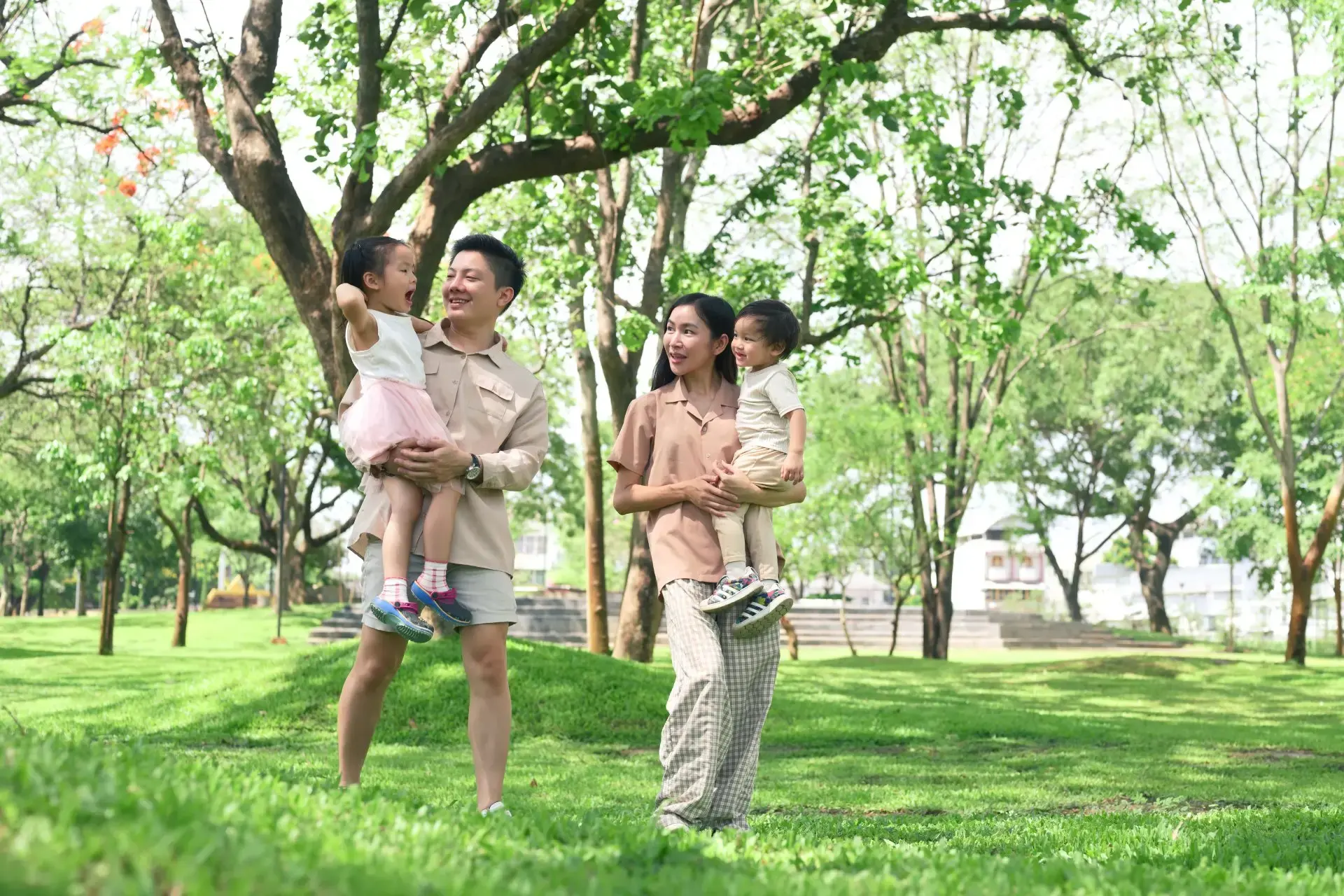 Asyanong tatay at nanay na buhat ang kanilang mga batang anak habang naglalakad sa parke at nagkukwentuhan.