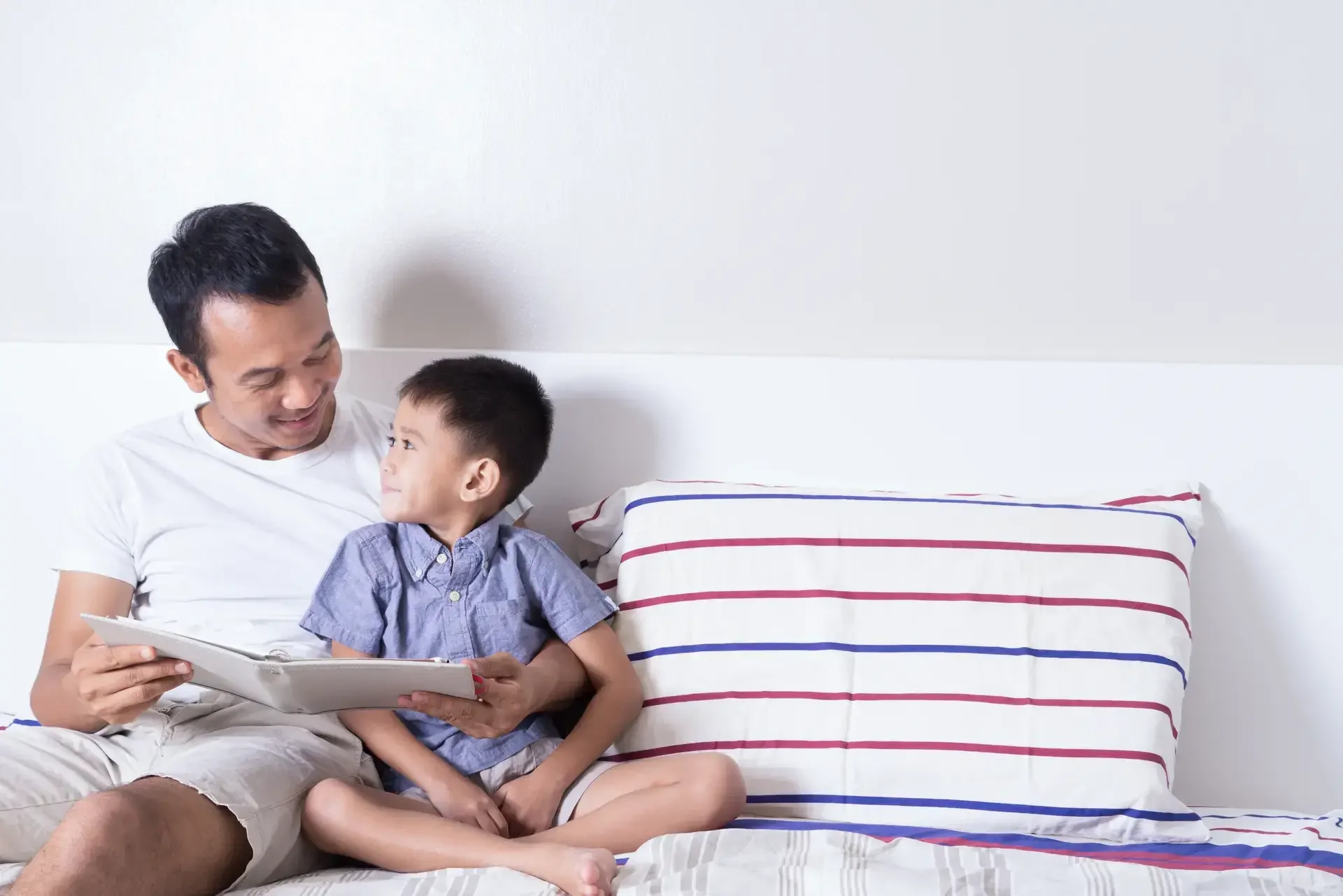 Masayang Asyanong tatay at anak na nagbabasa ng libro sa kama.