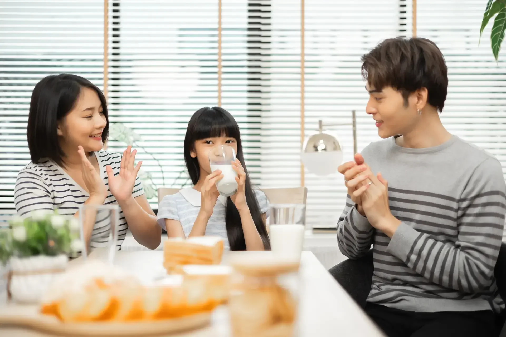 Masayang Asyanong pamilya na nasa kitchen habang ang batang anak na babae ay may hawak na baso ng gatas.