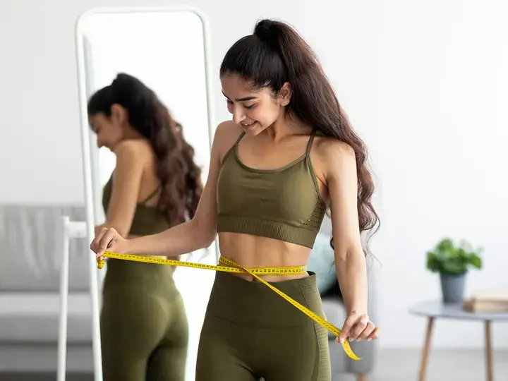 Fit Indian woman in sportswear measuring waist with tape near mirror